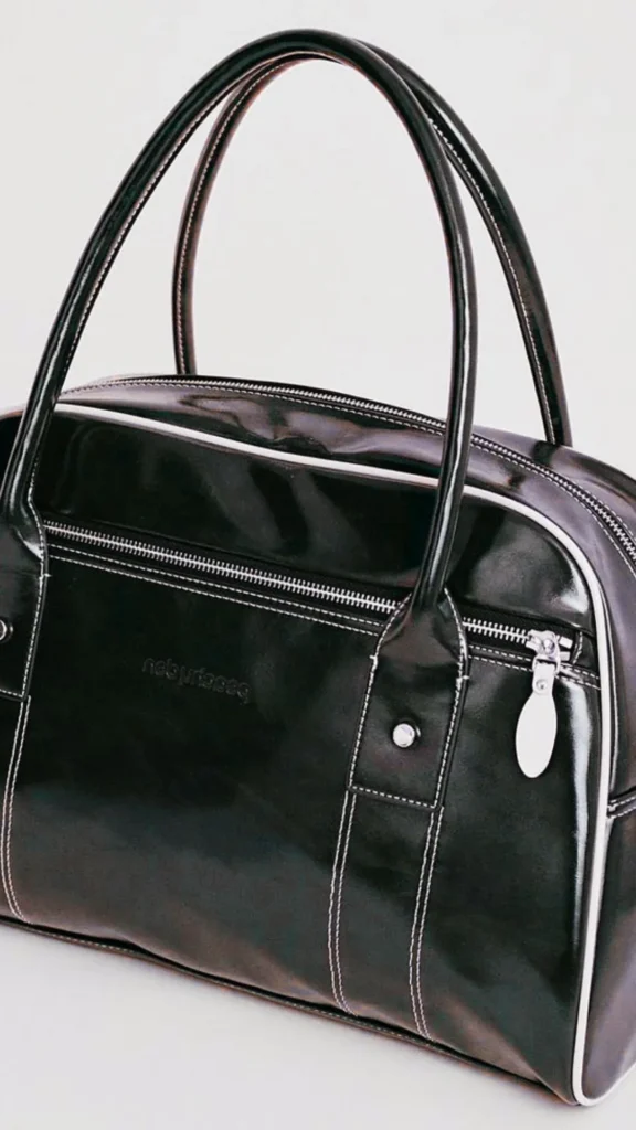 A close-up shot of a black glossy patent leather bowler-style handbag against a plain light gray background. The bag features structured top handles, white contrast stitching along the seams, a zippered front pocket with a silver metal pull, and white piping details along its curved edges.