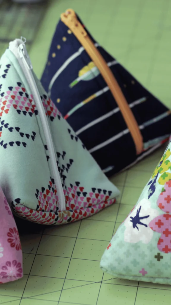 A close-up shot of multiple handmade fabric pouches sewn into unique, three-dimensional triangle pyramid shapes. One pouch features a light mint green fabric covered in small pink and black heart patterns with a white zipper. Another pouch in the background is dark navy blue with white stripes and a contrasting yellow-gold zipper. The completed projects are resting on a green self-healing cutting mat with grid lines.