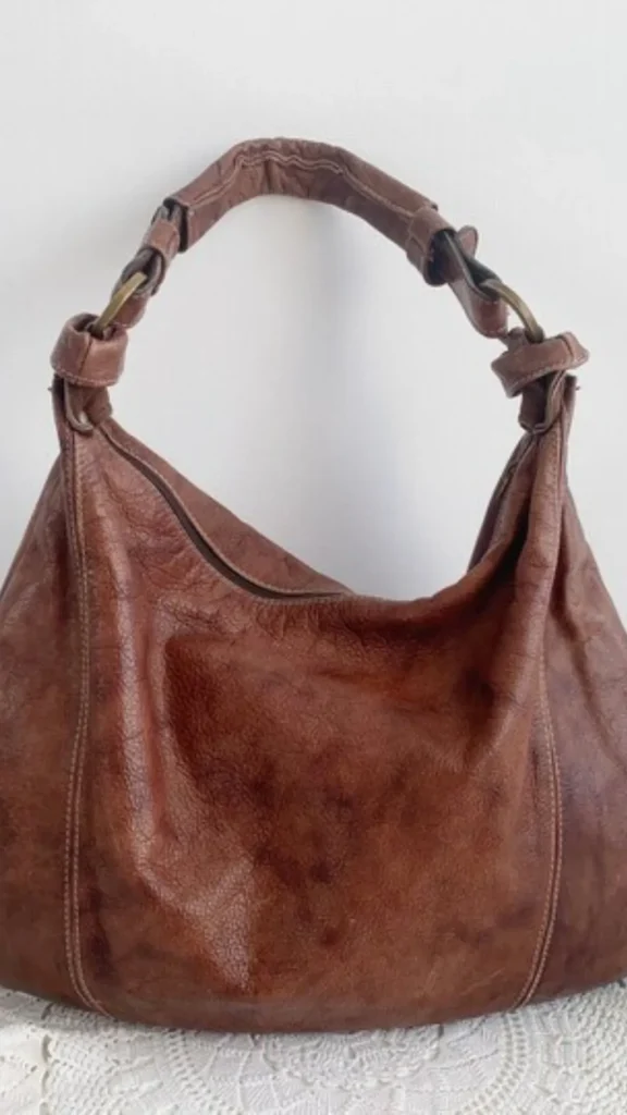 Front view of a slouchy, crescent-shaped brown leather hobo bag sitting on a white lace tablecloth against a plain white background. The bag is made from crinkled, soft tan-brown leather with vertical side seams. It features a matching wide leather shoulder strap connected by subtle antique brass rings and knotted leather details at the base of the handle. The unstructured bag dips naturally in the center, showing a hint of its top zipper line.