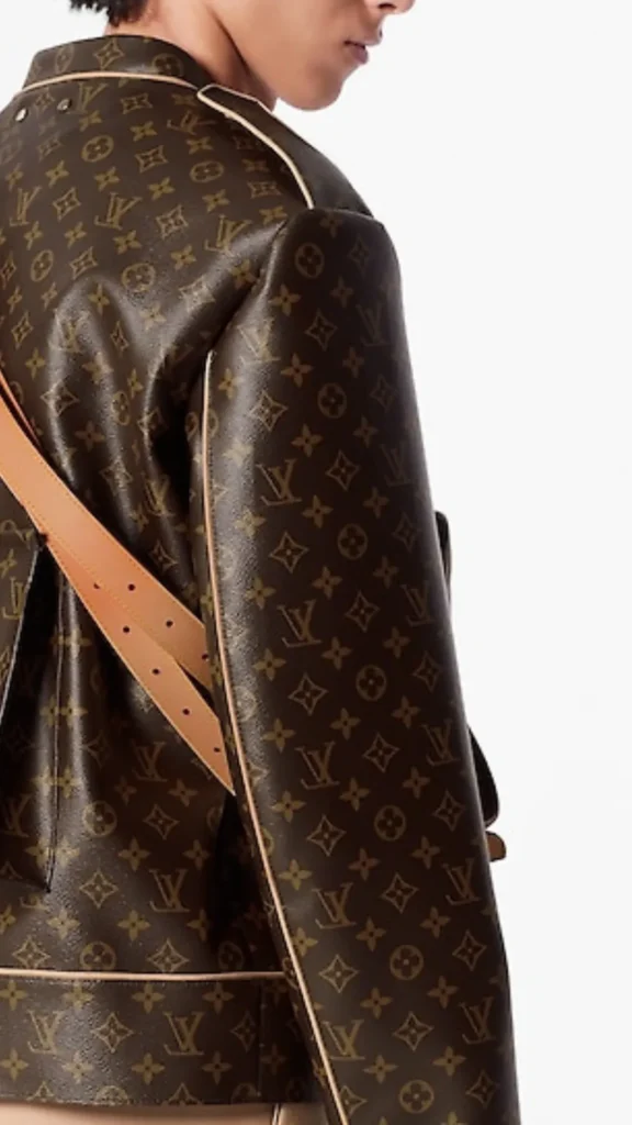 A close-up side profile shot of a model wearing a structured Louis Vuitton leather jacket featuring the iconic brown and tan LV monogram pattern all over. The jacket has light tan leather piping details along the seams and collar. A thick, adjustable tan leather shoulder strap is slung across the back of the jacket. The image is cropped to show the shoulder and arm details against a crisp, solid white background.