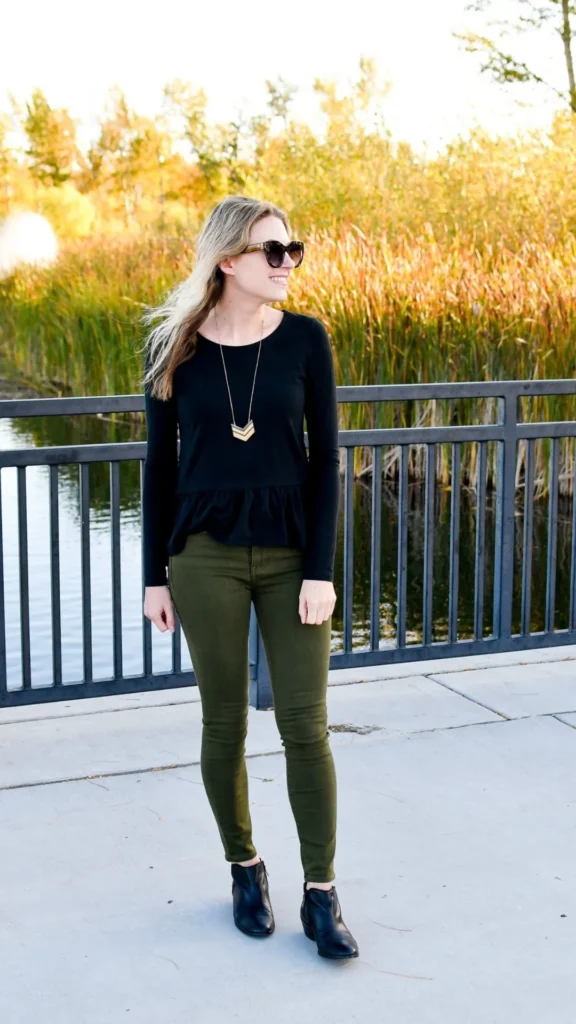 A smiling blonde woman wearing sunglasses stands outdoors on a concrete boardwalk by a metal railing with tall golden reeds in the background. She wears a black long-sleeve peplum top, olive green skinny pants, black leather ankle boots, and a long gold chevron pendant necklace. The sun creates a warm, autumnal light around her.