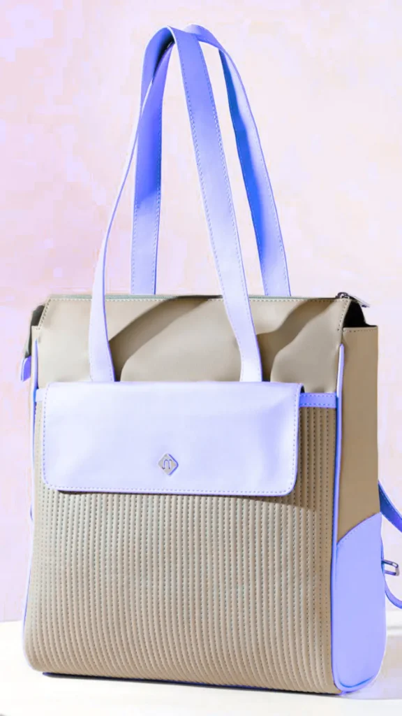 A structured, two-tone convertible tote bag standing upright against a soft pinkish-purple background. The bag features a textured, vertically quilted tan body with light lavender-purple accents, including a front flap pocket with a small silver diamond logo, purple trim, and long lavender shoulder straps.