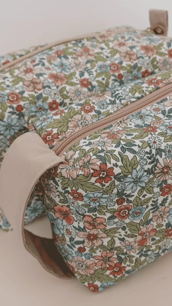 A close-up shot of a handmade, structured rectangular cosmetics pouch with boxed corners that allow it to sit upright. The bag is crafted from a vintage-style floral fabric featuring small pink, red, and blue flowers with green leaves. It has a matching dusty pink zipper running along the top and a wide, fabric loop handle attached to the side panel. The completed project is displayed against a clean, neutral background.