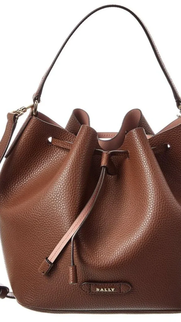 Close-up shot of a structured dark brown pebbled leather bucket bag against a clean white background. The bag features a cinched drawstring opening with thin matching leather ties and a smooth, arched top handle. A gold-toned metal clip connects a side shoulder strap. The interior lining is a light dusty pink color, visible through the folds of the cinched top. Near the bottom base, a small, stitched leather plaque displays the word "BALLY" in metallic gold lettering.
