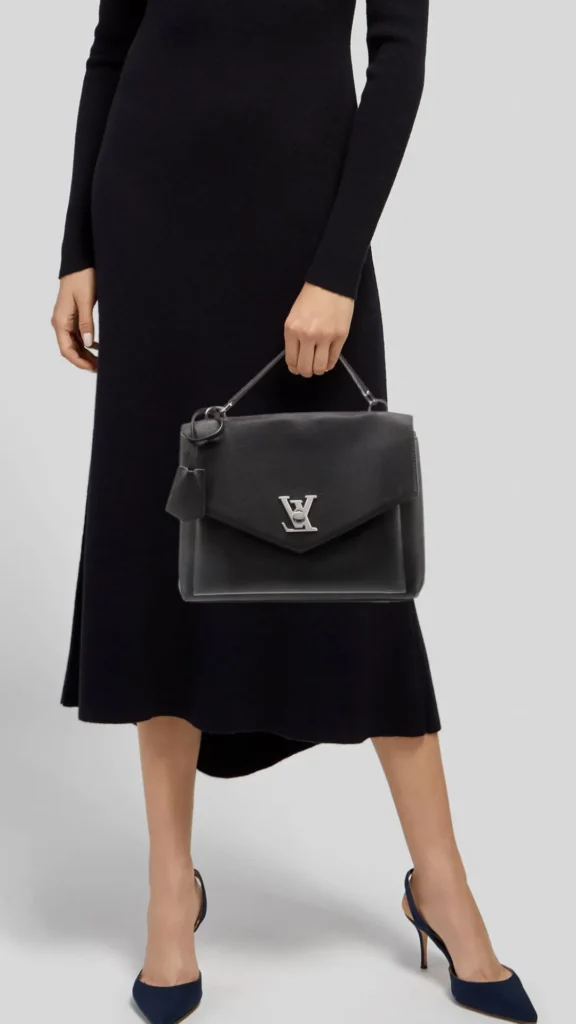 A studio portrait of a woman standing against a plain gray background. She is wearing a long-sleeved, knee-length knit black midi dress paired with dark navy blue pointed-toe stiletto heels. In her hand, she carries a structured black leather Louis Vuitton Lockme top-handle handbag, which features a prominent silver LV twist-lock logo on the front flap and a matching black leather clochette key tag hanging from the side.