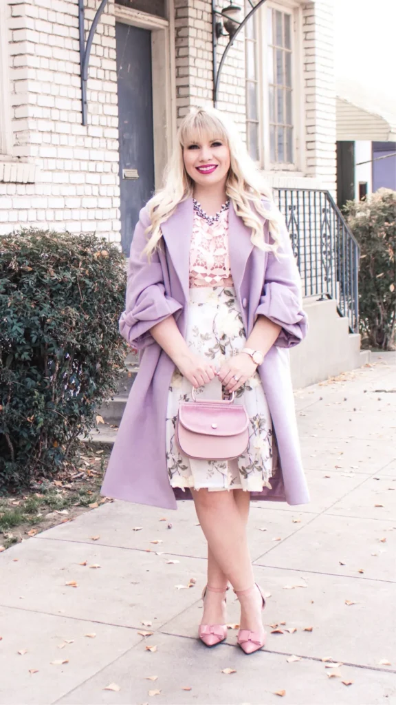 A smiling blonde woman with bangs stands outdoors on a sidewalk wearing a pastel outfit. She wears a peach lace top, a cream floral A-line skirt, and a mid-length lavender coat draped over her shoulders. She holds a small pink structured handbag in front of her with both hands and wears pink ankle-strap heels adorned with bows on the toes. A light brick building with a black door forms the background.