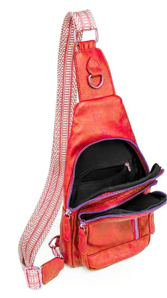 An upright, studio shot of an orange-red iridescent metallic sling crossbody backpack against a white background. The bag has multiple open zippered compartments showing a black fabric interior with pockets. It features purple accents along the zippers and a thick, adjustable woven strap with a geometric orange, white, and purple pattern.